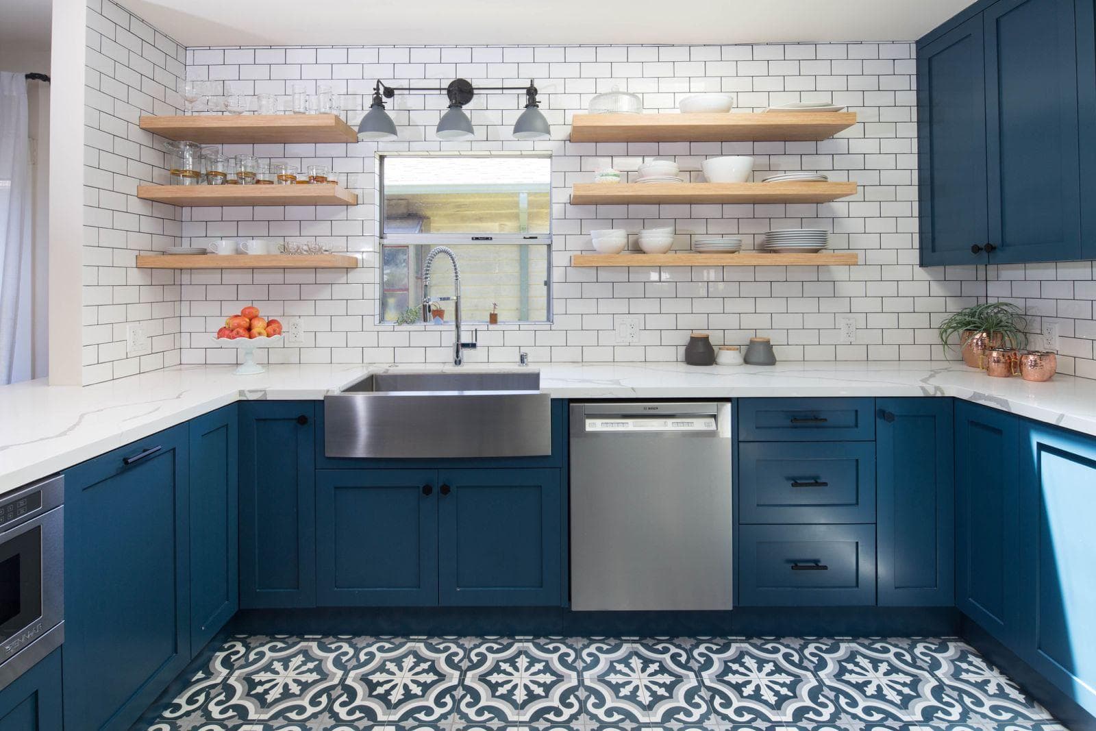 Blue shaker kitchen with white subway tile, open wood shelving, a stainless farmhouse sink, and patterned encaustic floor tile.